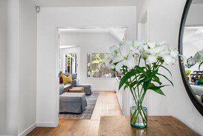 A hallway with a wooden floor, a glass vase with white flowers, a mirror, and a view into a living room with a sofa and cushions. - KaleidoNook | 3BR + Parking + Private Yard (Christchurch)