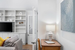A modern living room with a flat-screen TV, a sofa with cushions, a wooden table with a lamp, and a white shelving unit with decorative items.