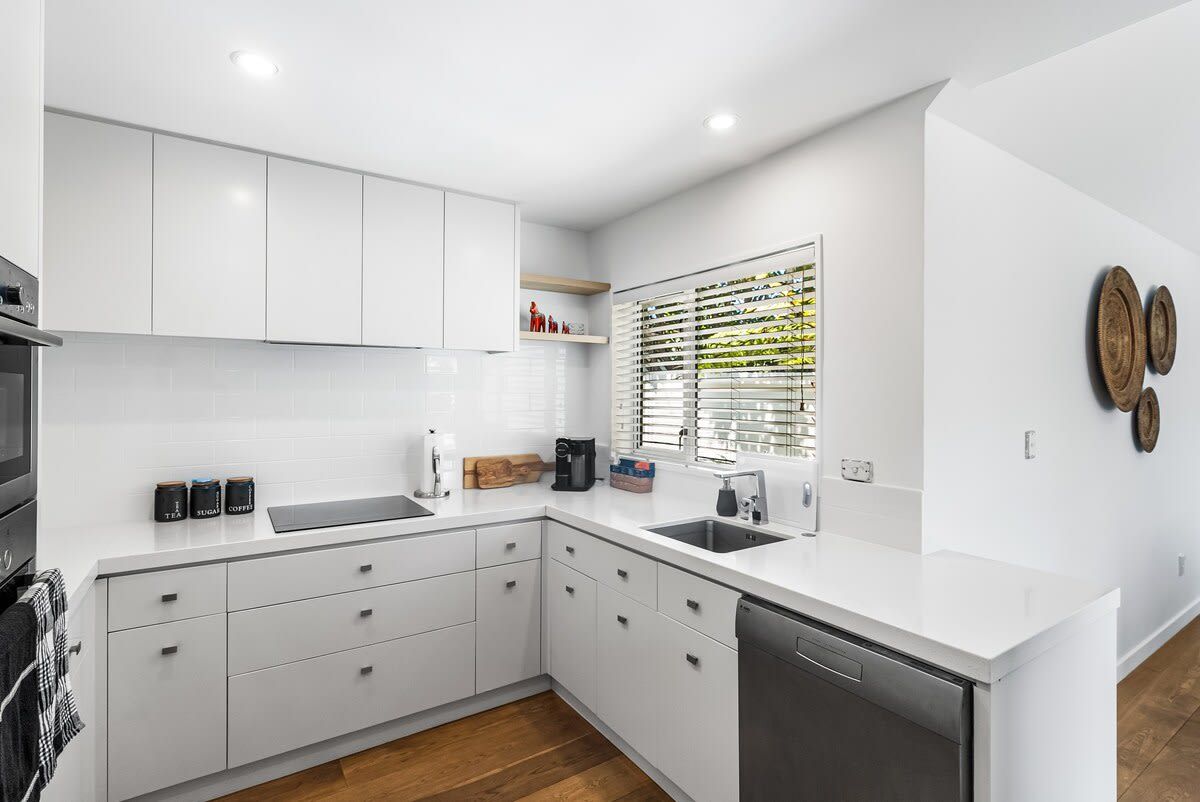 Una cocina moderna con armarios blancos, lavavajillas y una ventana con persianas.