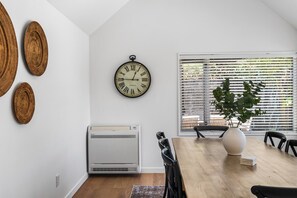 A dining area with a wooden table, chairs, a clock, and a window with blinds. - KaleidoNook | 3BR + Parking + Private Yard (Christchurch)