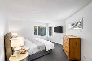 A modern bedroom with a bed, bedside lamp, dresser, TV, and windows.