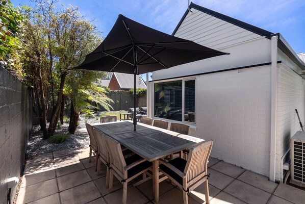 An outdoor patio with a wooden table and chairs, a black umbrella, and a white building with windows.