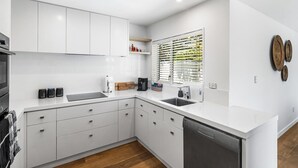 A modern kitchen with white cabinetry, a dishwasher, and a window with blinds. - KaleidoNook | 3BR + Parking + Private Yard (Christchurch)