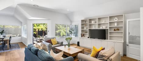 A spacious living room with a sofa, coffee table, and TV. There is a dining area with a table and chairs. The room has large windows and a view of greenery outside.