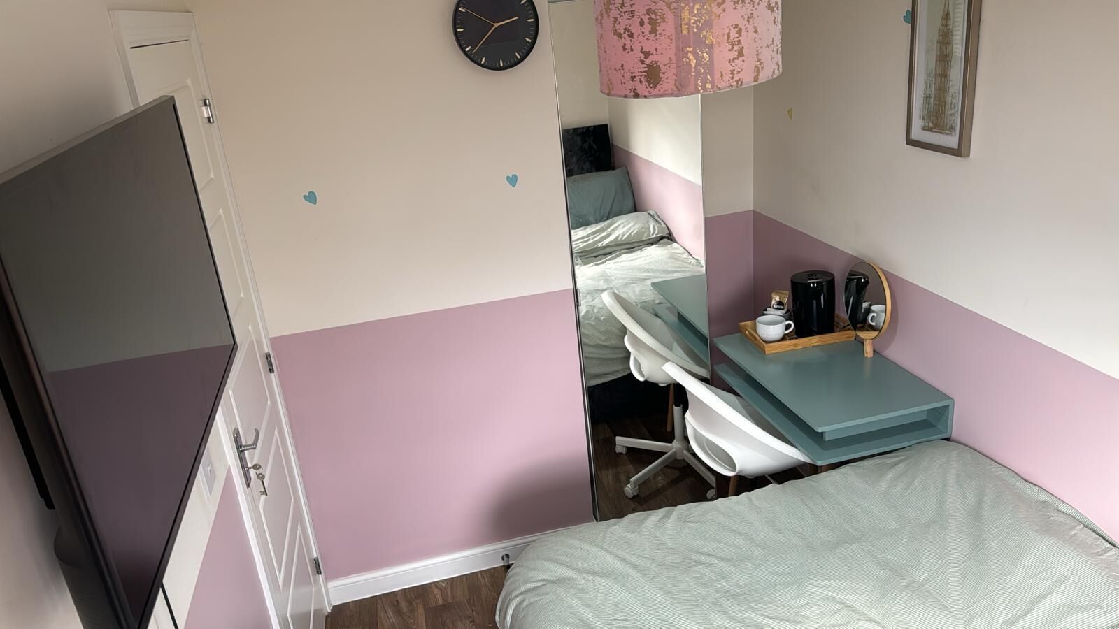 A bedroom with a bed, a desk with a chair, a clock, and a coffee maker.