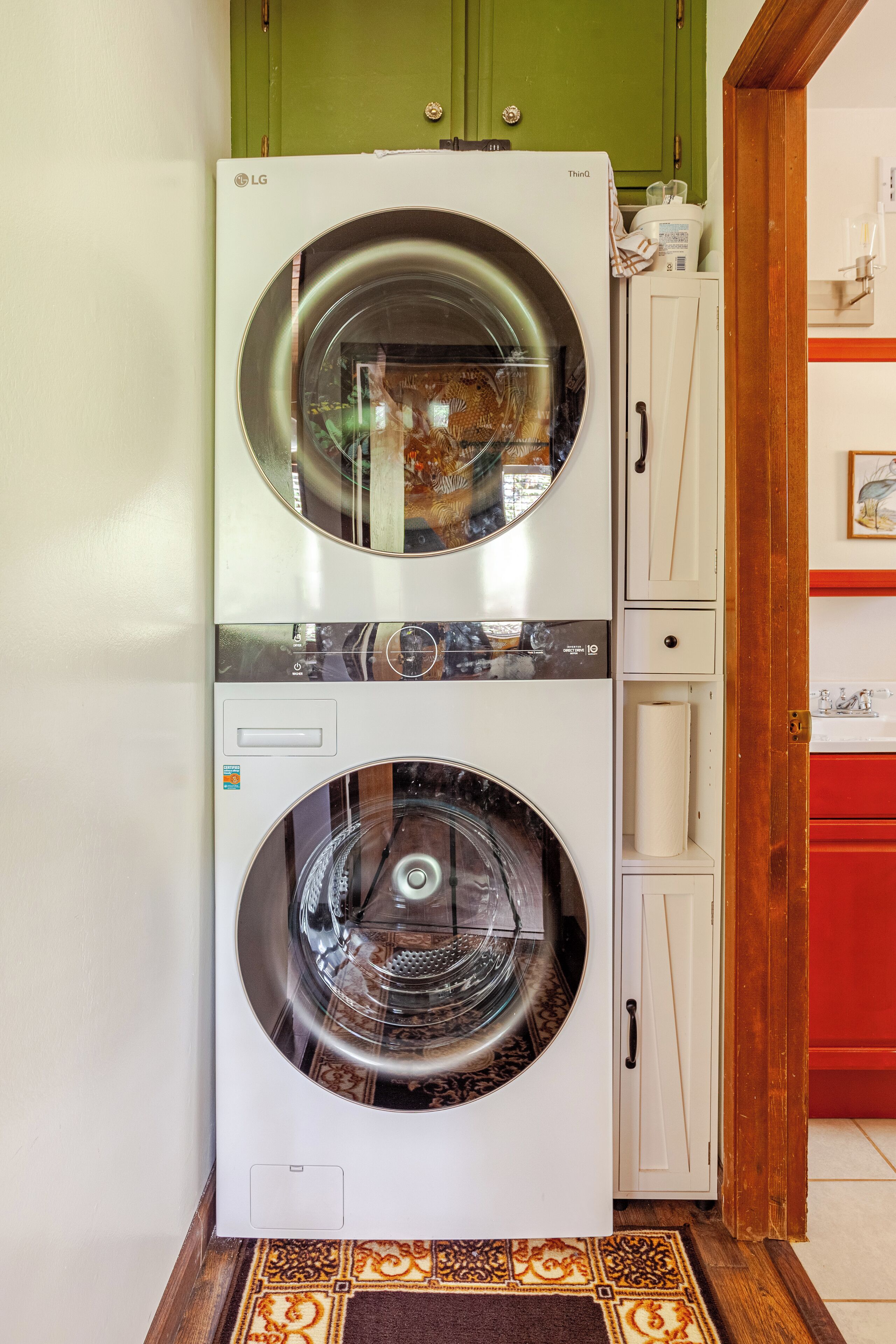 A stacked washer and dryer unit in a laundry room with green cabinets and a patterned rug.