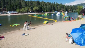 Een strand met een uitkijktoren voor badmeesters, speeltoestellen in het water en een bebost heuvel op de achtergrond.