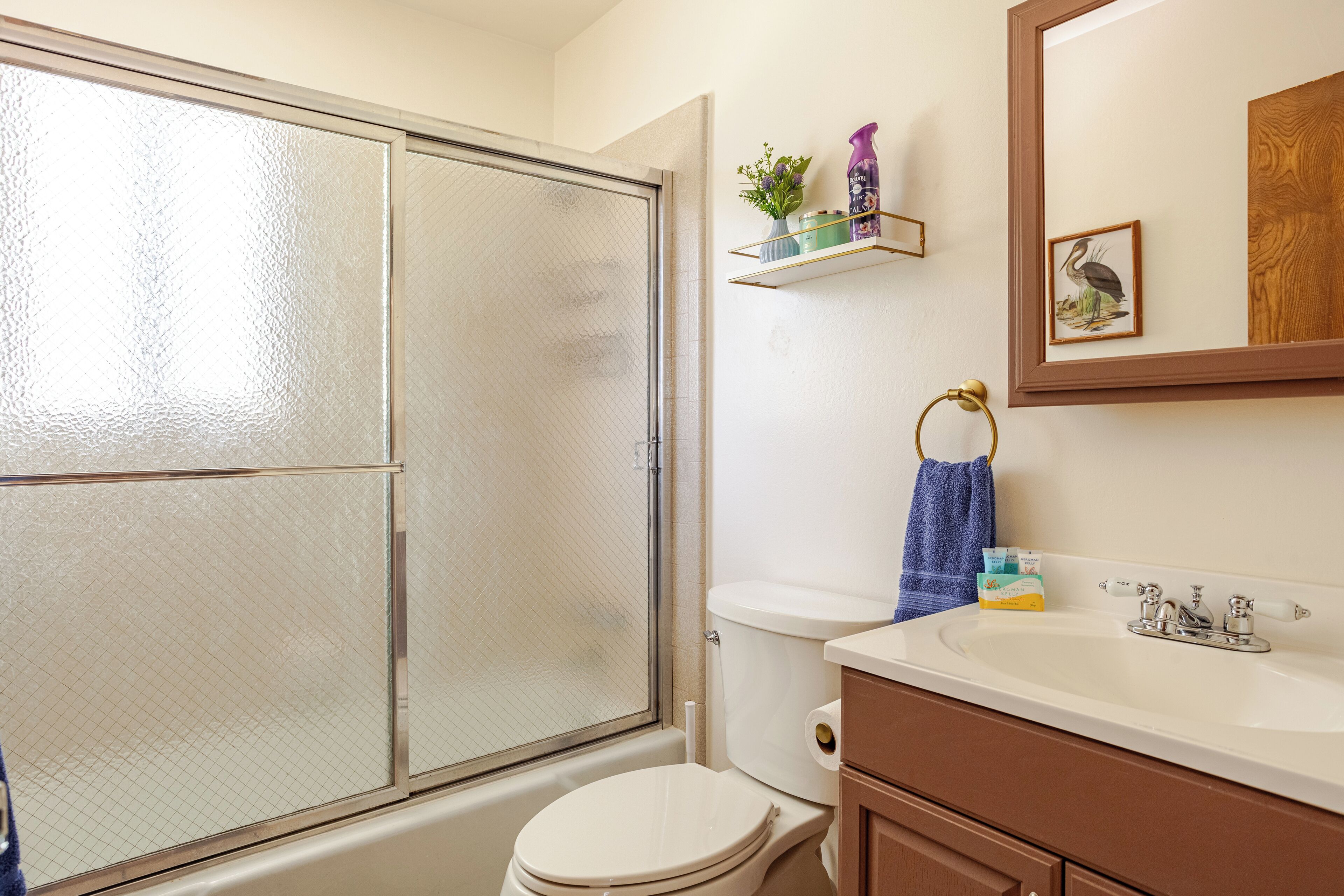 A bathroom with a glass shower door, a toilet, a sink with a mirror, and a shelf with toiletries.