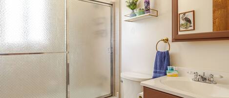 A bathroom with a glass shower door, a toilet, a sink with a mirror, and a shelf with toiletries.