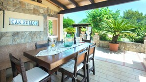 Un patio al aire libre con un muro de piedra, una mesa de madera con vasos y una jarra, y un cartel que dice "CAN FULLOS".