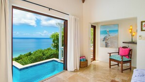 A room with a view of the ocean, a pool, a chair with a cushion, and a painting on the wall.