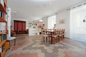 A room with a hexagonal patterned floor, a dining table with chairs, a bookshelf, and a large window with sheer curtains.