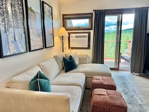 A living room with a sectional sofa, a standing heater, and a view of a deck.