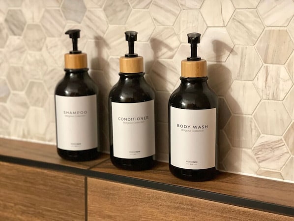 Trois bouteilles de gel douche, d’après-shampoing et de shampoing sont posées sur une étagère en bois, devant un mur recouvert de carreaux hexagonaux.