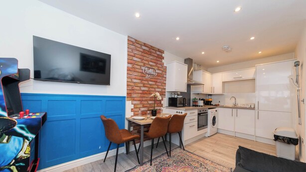 A modern kitchen with white cabinets, a brick accent wall, a flat-screen TV, and a dining area with chairs and a table.