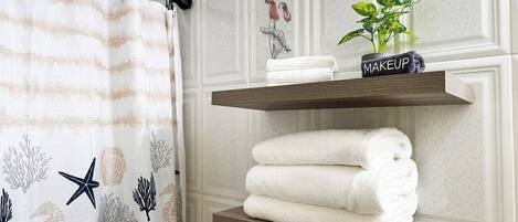 Un baño con cortina de ducha decorada con motivos marinos, un estante con toallas dobladas y una pequeña planta etiquetada 'MAKEUP' sobre una repisa de madera.