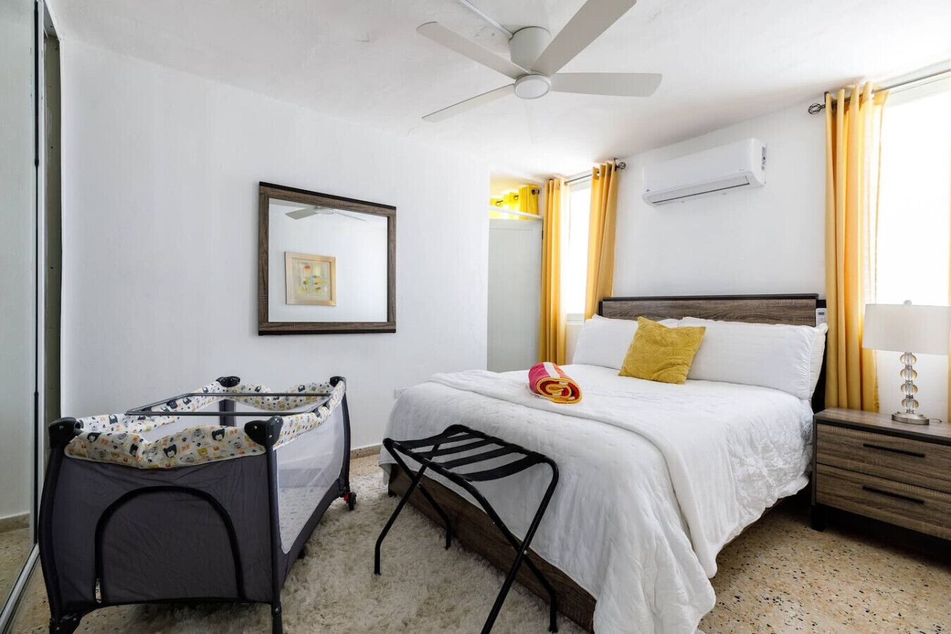 A hotel room with a bed, a crib, a ceiling fan, a bedside table with a lamp, and a window with curtains.