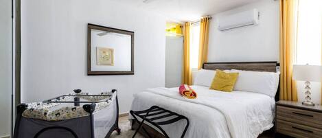 A hotel room with a bed, a crib, a ceiling fan, a bedside table with a lamp, and a window with curtains.