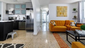 Eine moderne Küche mit Edelstahlgeräten, ein Essbereich mit einem gelben Sofa und ein Wohnzimmer mit Deckenventilator.