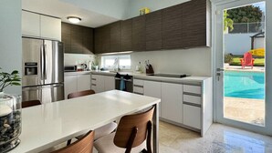 A modern kitchen with a built-in refrigerator, a dining table with chairs, and a view of a swimming pool through an open door.