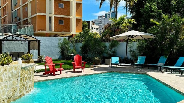 A swimming pool with red chairs and umbrellas, surrounded by greenery and buildings.