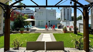 A covered outdoor area with a pool, red chairs, and a view of a white building in the background.