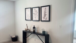 Une pièce avec une console noire adossée à un mur blanc, trois reproductions botaniques encadrées au-dessus, un petit panier et une lampe sur pied.