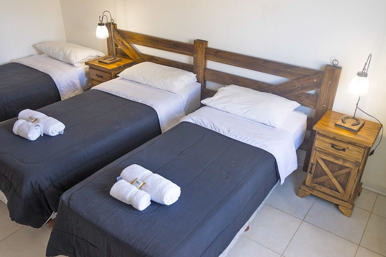 A room with three single beds, each with a wooden headboard, white pillows, and a dark grey bedspread. There are wooden nightstands with lamps and a small wooden cabinet with a hook.