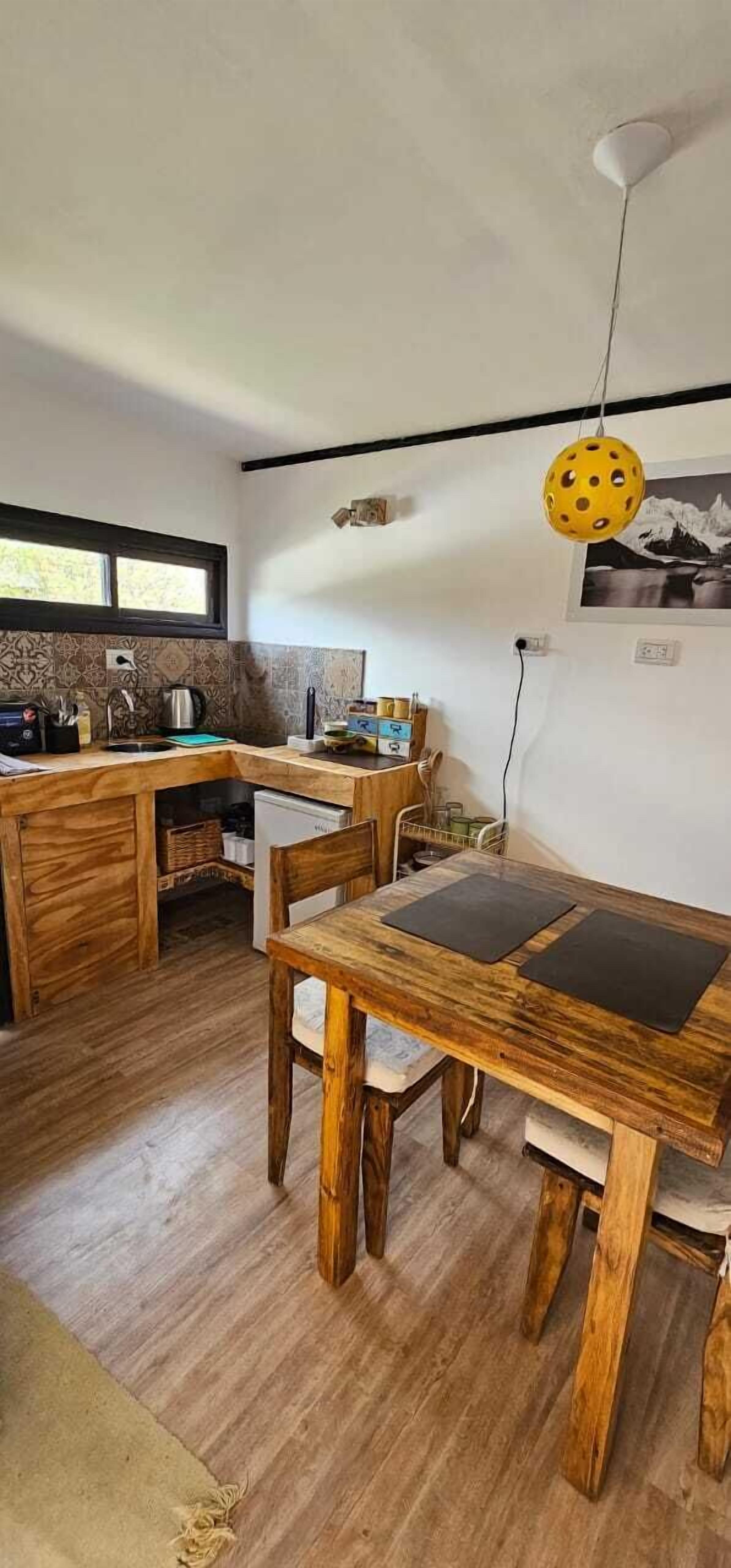 A compact kitchen with wooden cabinetry, a dining table with chairs, a hanging yellow light fixture, and a window providing natural light.