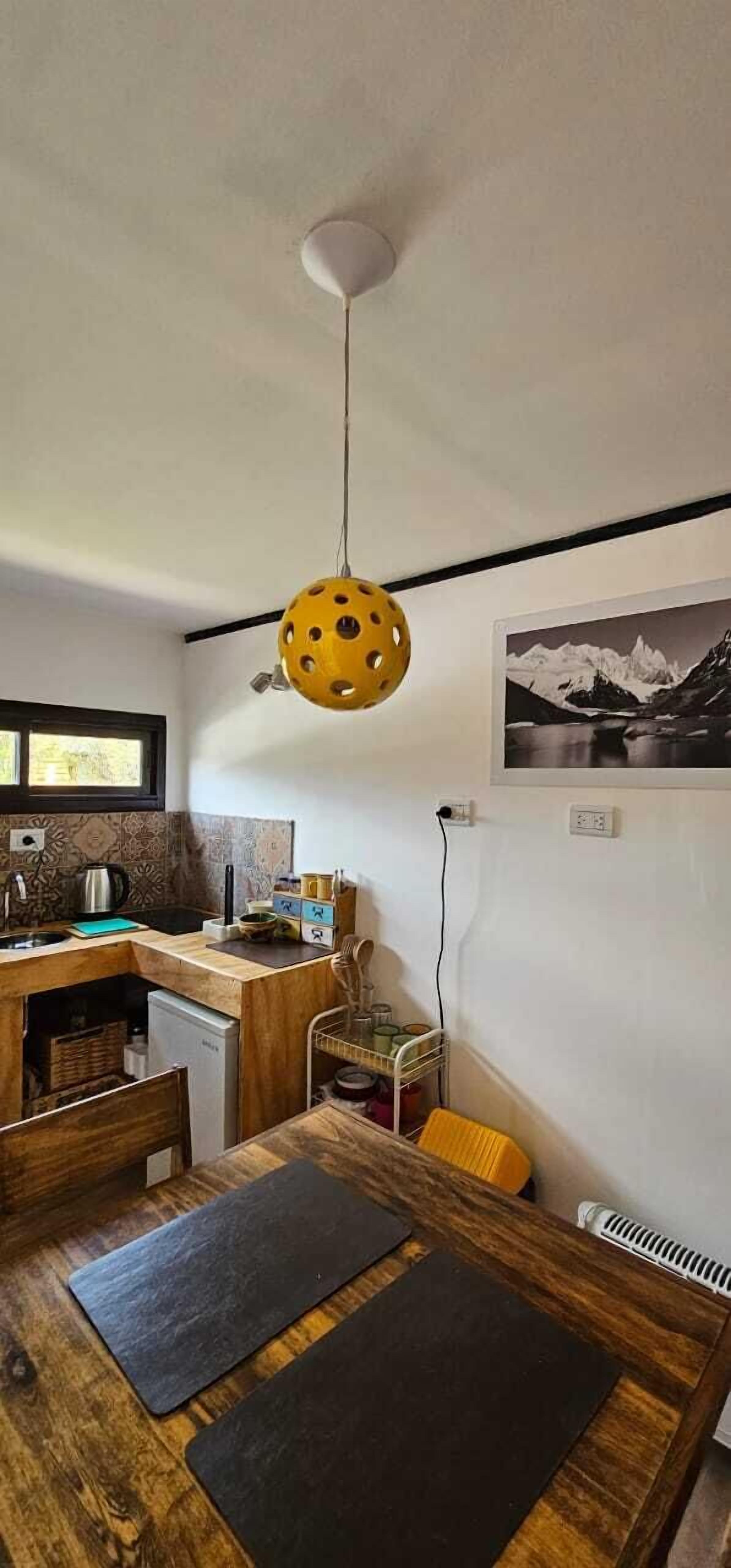 A compact kitchen with a wooden table, a yellow hanging light fixture, and a mountain landscape photo on the wall.