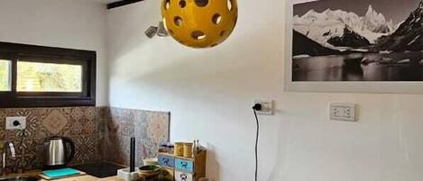 A compact kitchen with a wooden table, a yellow hanging light fixture, and a mountain landscape photo on the wall.