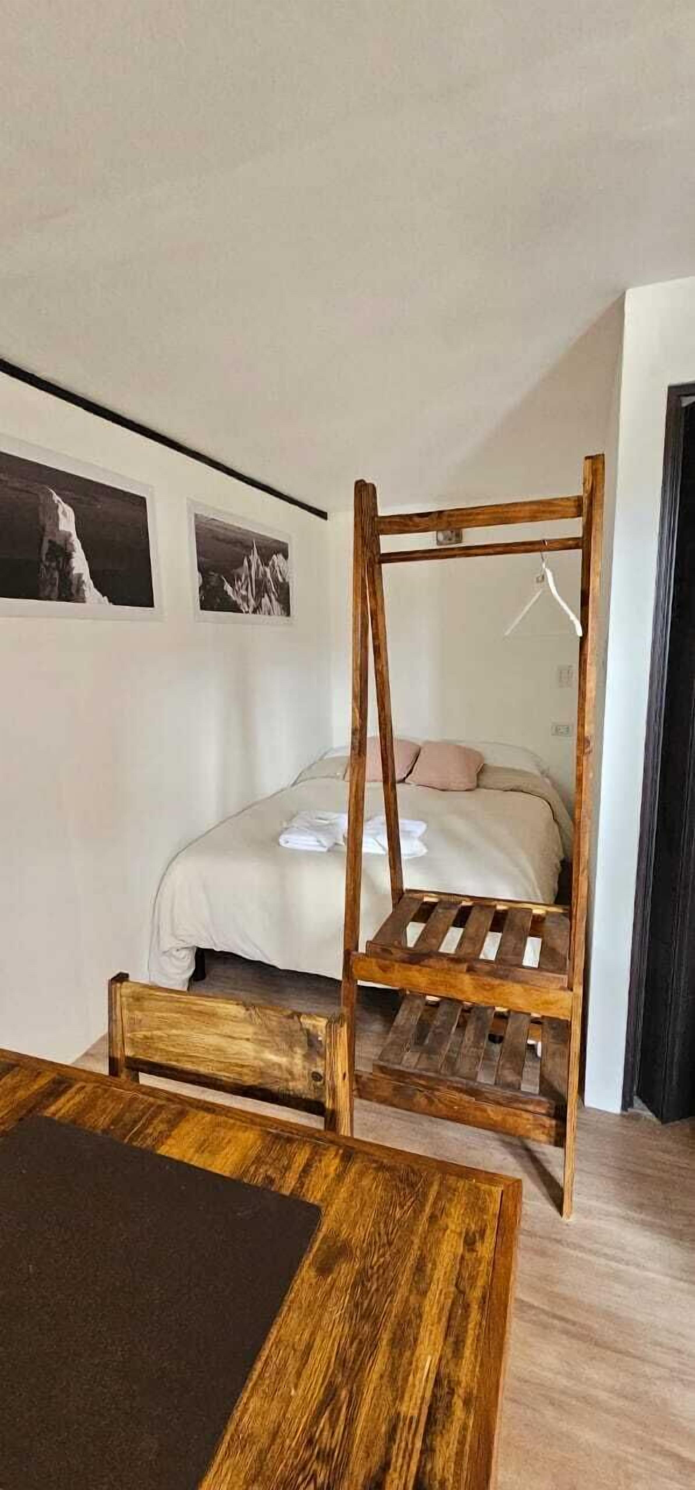 Una habitación con estructura de cama de madera, una mesita de madera y cama con colchón blanco y ropa de cama color crema.