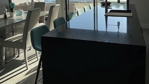 Een moderne eetruimte met een grote glazen tafel en stoelen, grote ramen met uitzicht op een meer en bomen, en een plafondventilator.