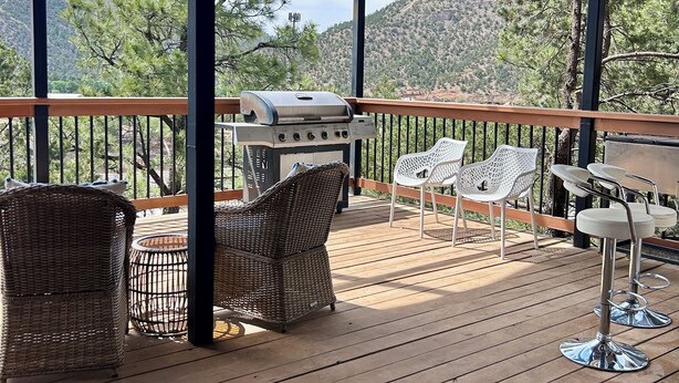 Une terrasse extérieure couverte, équipée d’un barbecue, de fauteuils en osier et de chaises en plastique blanc.