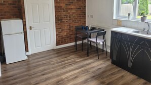 A compact kitchen with a brick wall, a white door, a refrigerator, a dining table with chairs, and a window with a view of greenery outside. - 1 Bedroom - Sleeps 3 - Free Parking (Luton)