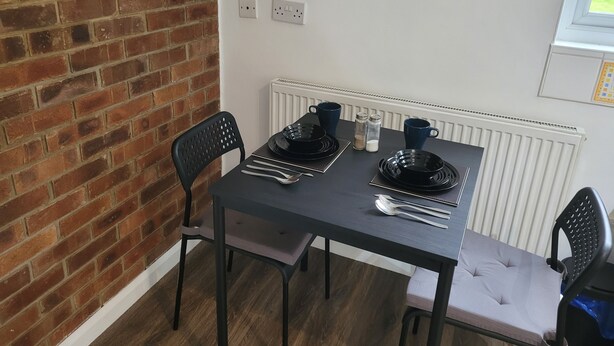 A dining area with a brick wall, a table set with black plates, silverware, and blue cups, and a window with blinds. - 1 Bedroom - Sleeps 3 - Free Parking (Luton)