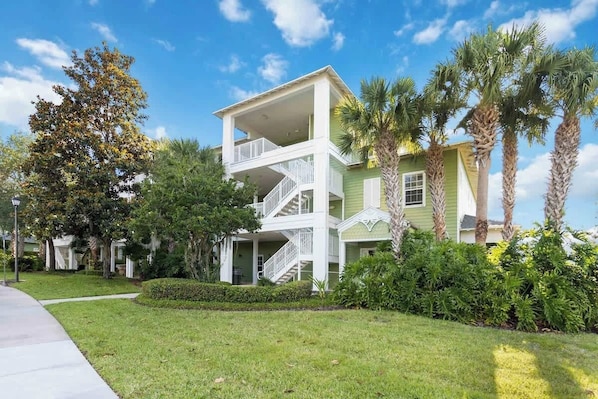 A multi-story building with a balcony, surrounded by greenery and palm trees.