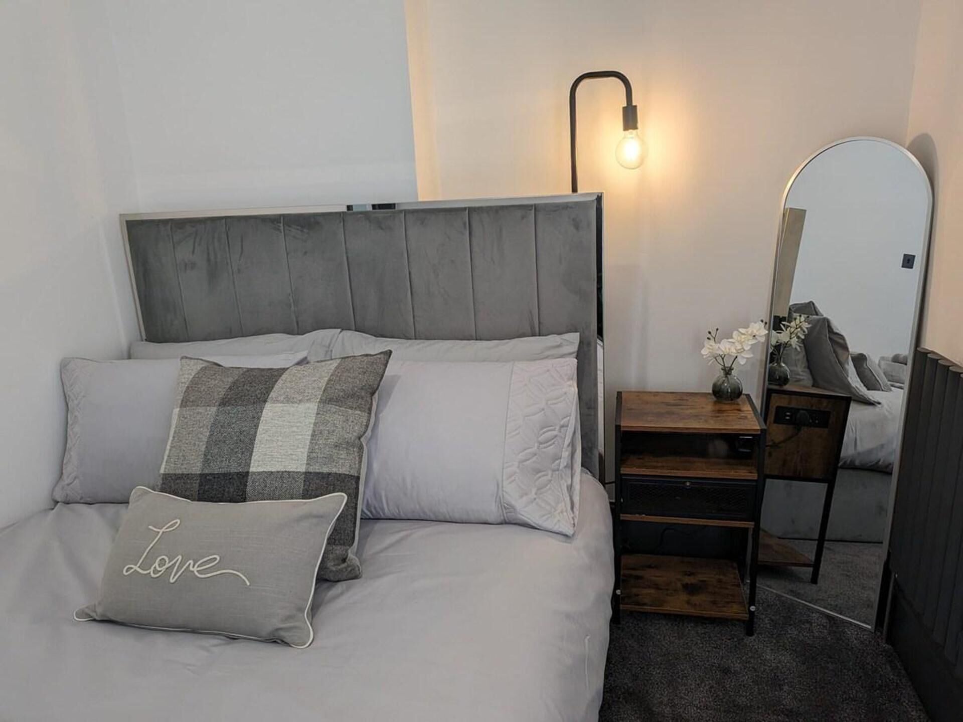 A modern bedroom with a grey upholstered headboard, a bedside table with a lamp, and a mirror.