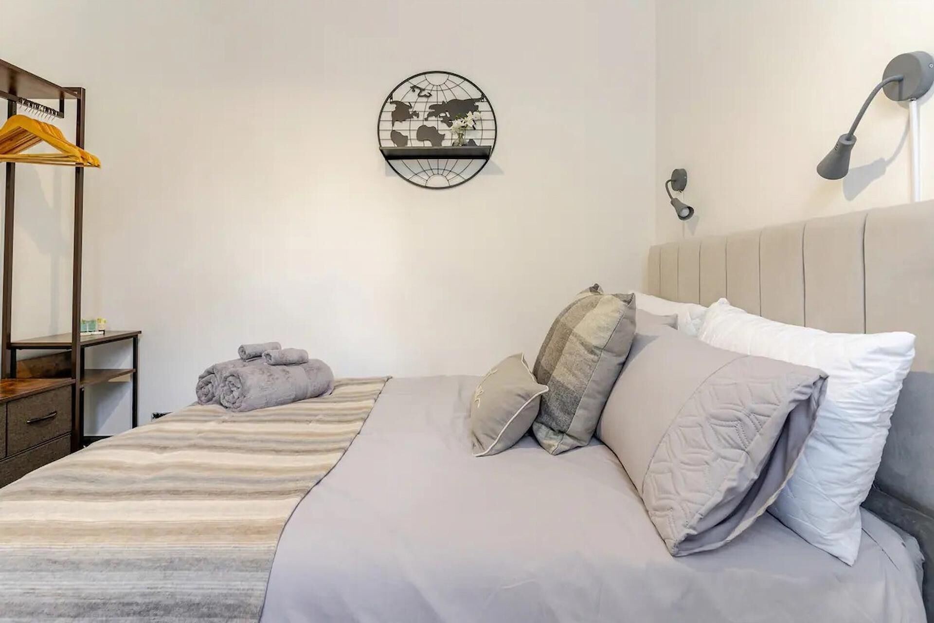 A neatly made bed with a striped bedspread, a world map artwork on the wall, a hanging rack with a yellow garment, and a bedside lamp.