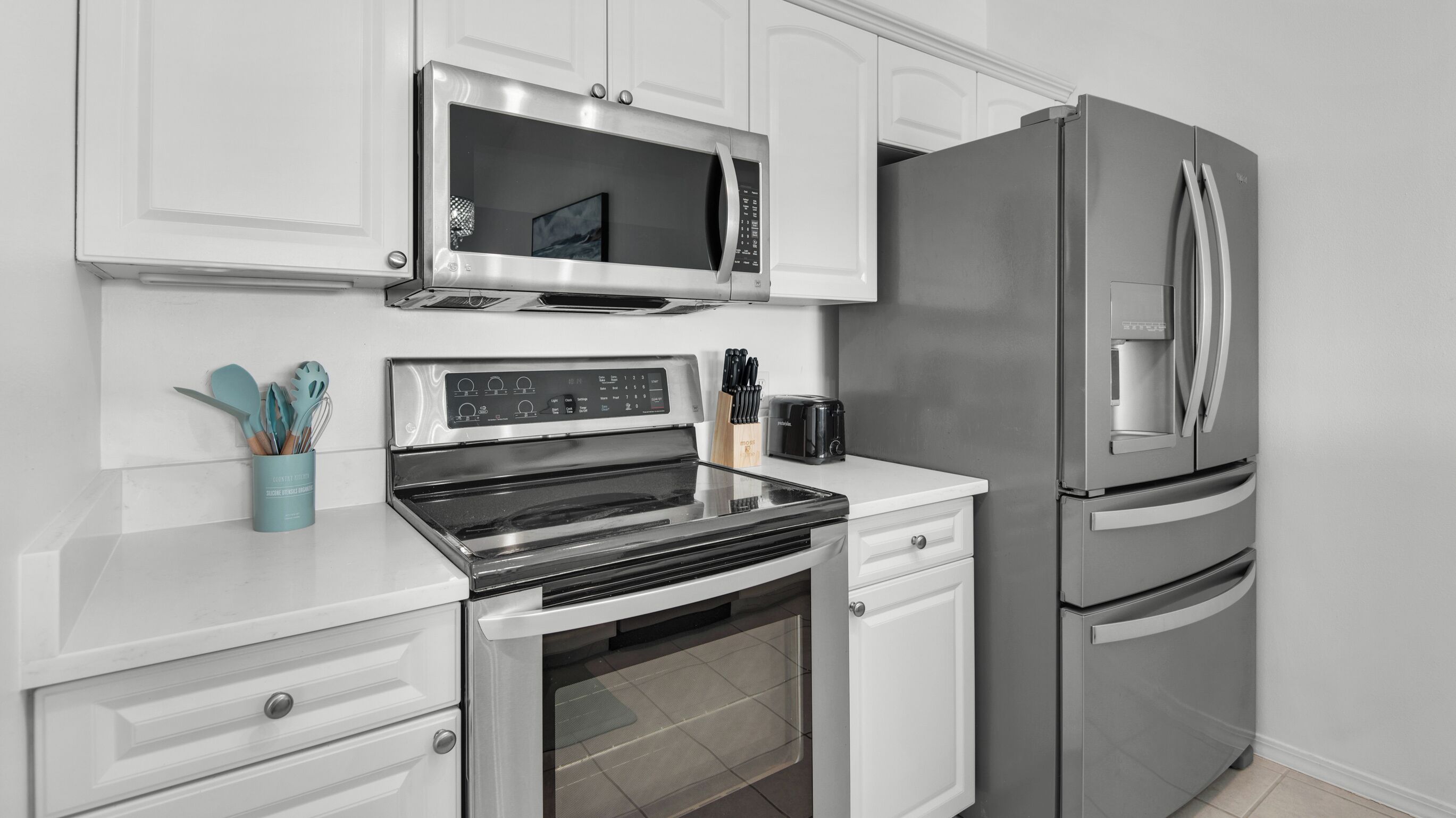 A modern kitchen with stainless steel appliances, including a microwave, oven, and refrigerator. There are white cabinets and a countertop with a toaster and a knife block.