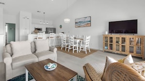 Ein modernes Wohnzimmer mit einer Couch, einem Couchtisch und einem Korbstuhl. Ein Essbereich mit weißen Stühlen und eine Holzvitrine mit Glastüren sind ebenfalls vorhanden. Ein Flachbildfernseher ist an der Wand montiert.