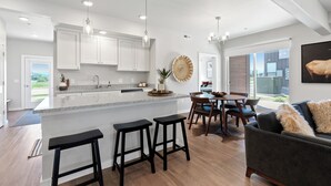 A modern kitchen with white cabinets, a granite countertop, and black barstools. There is a dining area with a table and chairs, and a large window offering a view of the outdoors. - Chic Bozeman Getaway - Minutes from Downtown! (Bozeman)