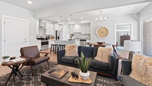 A modern living room with a sofa, coffee table, and decorative items. A kitchen is visible in the background with white cabinets and stainless steel appliances. - Chic Bozeman Getaway - Minutes from Downtown! (Bozeman)
