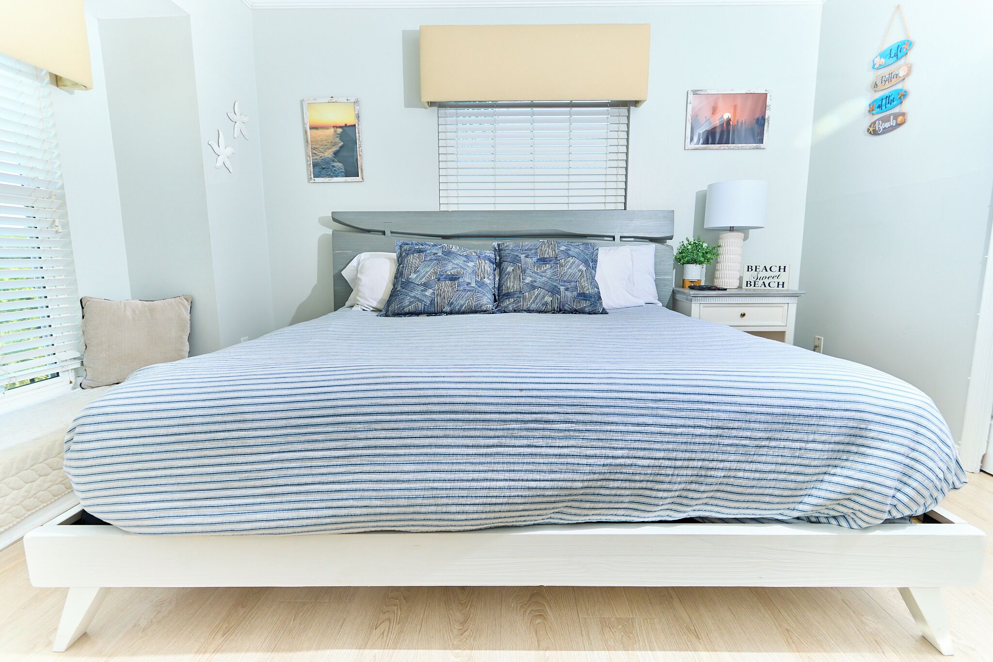 Ein ordentlich bezogenes Bett mit einer gestreiften Tagesdecke, ein Nachttisch mit Lampe und Wanddekorationen, darunter ein Schild mit Strandmotiv.