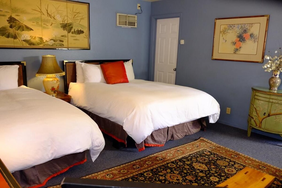 A hotel room with two beds, a nightstand, a lamp, a door, and a wall with a painting.