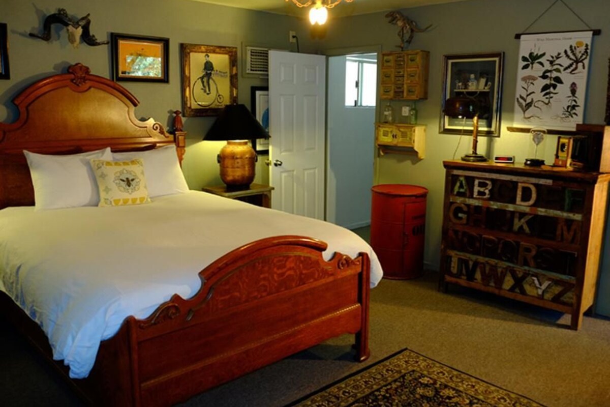 A bedroom with a wooden bed, a nightstand, a lamp, a red trash bin, a bookshelf with alphabet letters, and a wall with framed pictures.