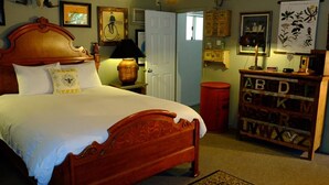 A bedroom with a wooden bed, a nightstand, a lamp, a red trash bin, a bookshelf with alphabet letters, and a wall with framed pictures.