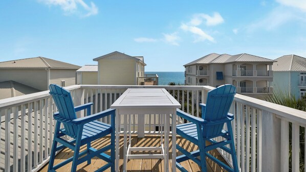 A balcony with two blue Adirondack chairs and a white table, overlooking a beach with buildings in the background. - 2 Minutes to Beach & Next to Pool - Free Beach Setup, Sleeps 12 (Santa Rosa Beach)