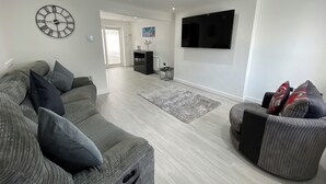 Un salon moderne avec un canapé d’angle gris, un fauteuil gris, un téléviseur à écran plat fixé au mur et une grande horloge murale.