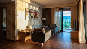 Un salon moderne avec un ensemble de canapés, une table basse et un téléviseur. Une grande fenêtre offre une vue sur la verdure et une montagne.
